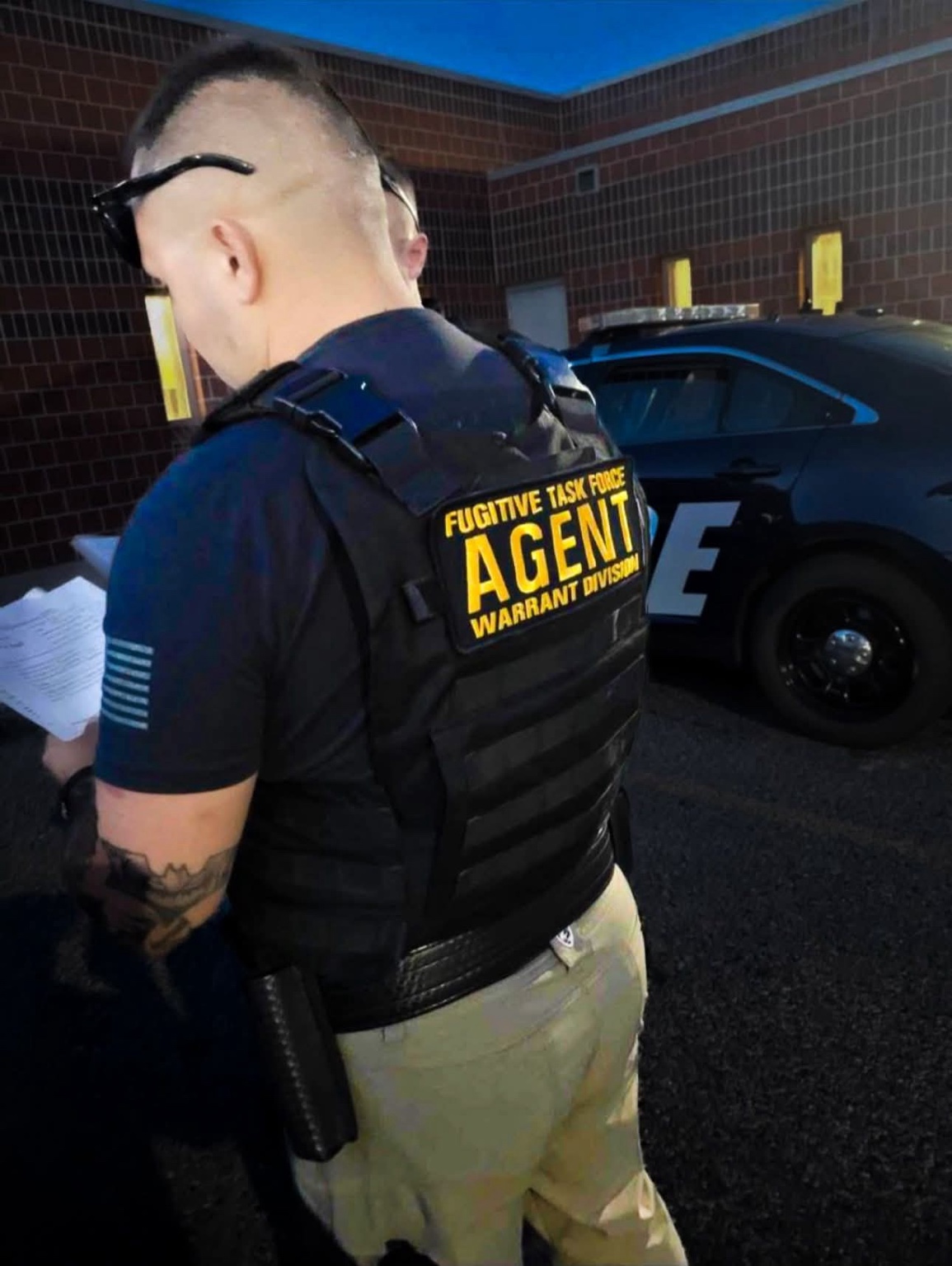 A fugitive recovery agent in a black vest reviewing paperwork beside an official vehicle outside a Montana facility.
